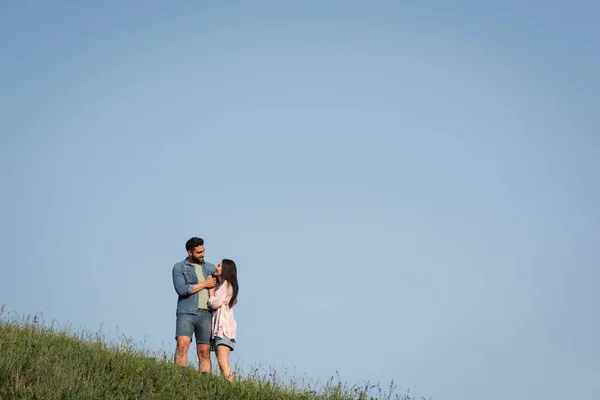 Romantic couple looking at each other and embracing in field under blue sky - foto de stock