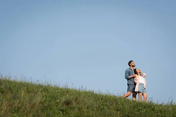 Happy couple in denim shorts standing on green hill under blue sky and looking away — Fotografia de Stock