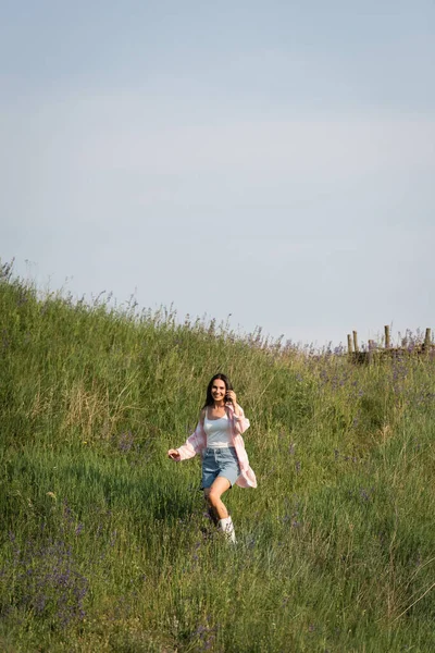 Cheerful brunette woman looking at camera in green meadow with wildflowers — Stockfoto