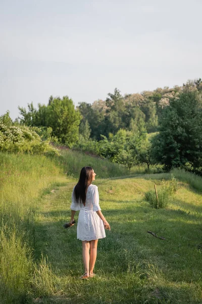 Visão traseira da mulher morena em vestido branco em pé na estrada rural perto da floresta — Fotografia de Stock