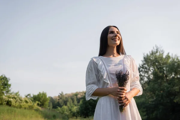Low angle view of happy woman in white dress holding lavender flowers outdoors — Stock Photo