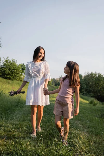 Feliz madre e hija tomados de la mano mientras caminan por un camino rústico en el día de verano - foto de stock