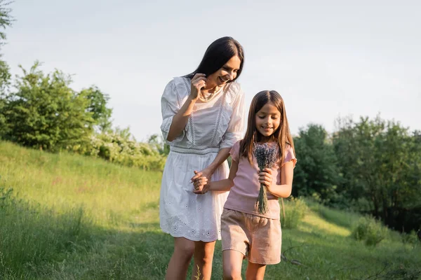 Lächelndes Mädchen mit Lavendelstrauß, Händchen haltend mit zufriedener Mutter auf dem Land — Stockfoto