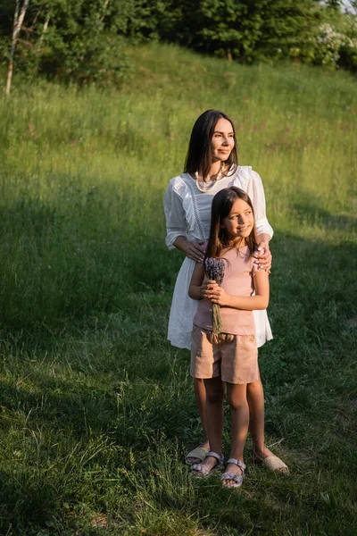 Smiling girl with lavender bouquet looking away near mom in countryside — Photo de stock