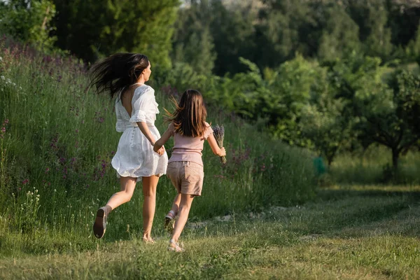 Back view of mother and child holding hands and running on grassy path towards forest — Stock Photo