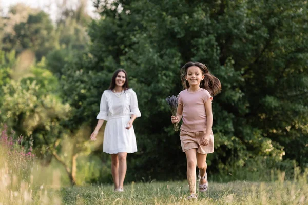 Full length of cheerful girl with lavender bouquet running near mom on blurred background — Fotografia de Stock