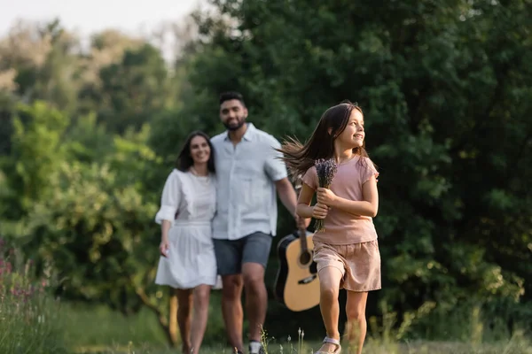 Blurred couple near joyful girl running with lavender flowers on summer day - foto de stock