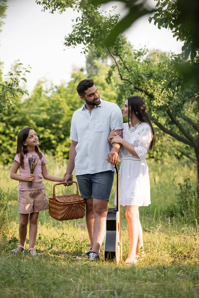 Happy family with wicker basket and acoustic guitar looking at each other in countryside garden — Stock Photo
