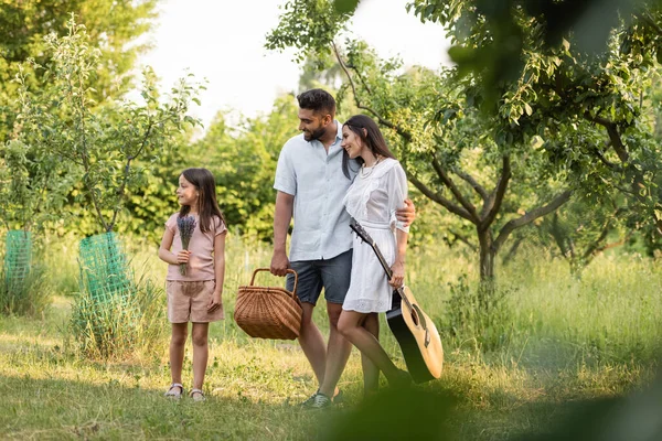 Couple heureux avec guitare acoustique et panier en osier regardant fille tenant des fleurs — Photo de stock