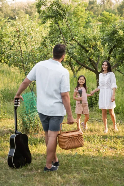 Hombre con cesta de mimbre y guitarra acústica cerca de esposa e hija sonriendo en el jardín - foto de stock