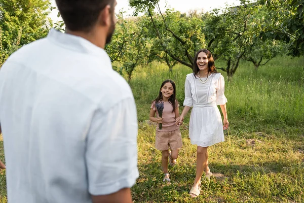 Blurred man near happy wife and daughter holding hands near green trees — Stockfoto