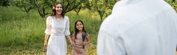 Joyful mother and daughter holding hands and smiling near blurred man outdoors, banner — Stock Photo