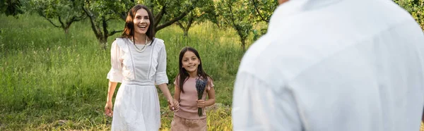 Freudige Mutter und Tochter Händchen haltend und lächelnd neben verschwommenem Mann im Freien, Banner — Stockfoto