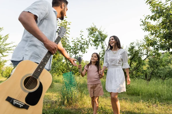 Man with guitar outstretching hand near wife and daughter smiling outdoors — Stock Photo