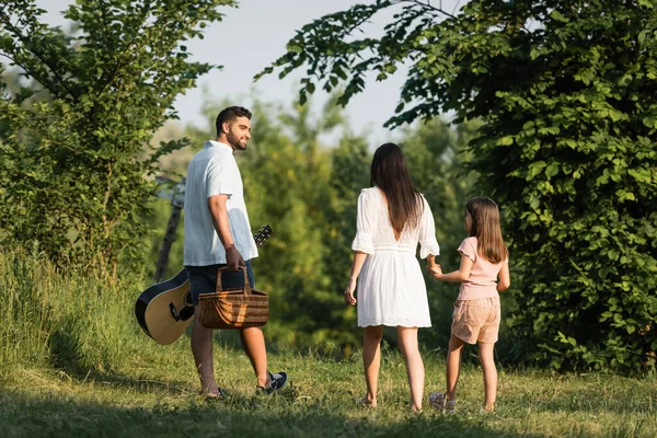 Man with guitar and wicker basket smiling near family walking in countryside — Stock Photo