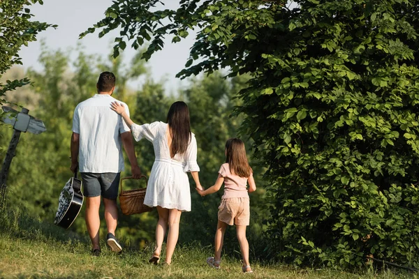 Back view of man with wicker basket and acoustic guitar walking with family outdoors - foto de stock