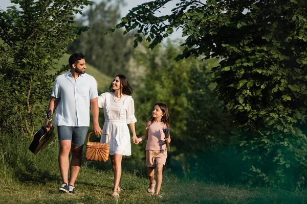 Comprimento total do homem feliz com guitarra acústica e cesta de vime andando com a família ao ar livre — Fotografia de Stock