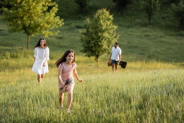 Fille heureuse avec bouquet courir dans le champ vert près des parents marchant sur fond flou — Photo de stock