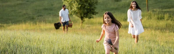 Cheerful child running in grassy meadow near parents on blurred background, banner - foto de stock