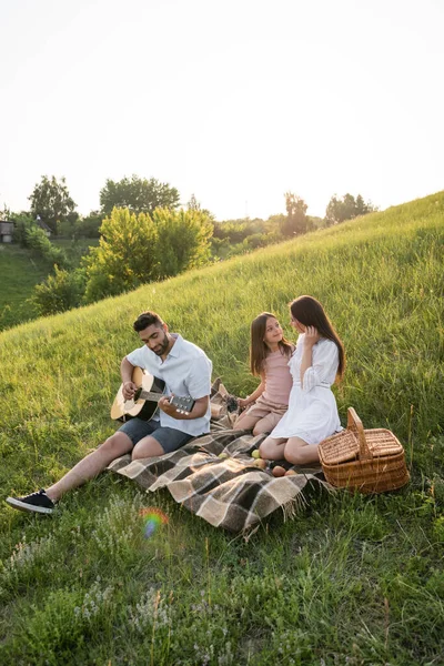 Homem tocando guitarra perto da família sentado no cobertor xadrez no prado verde — Fotografia de Stock