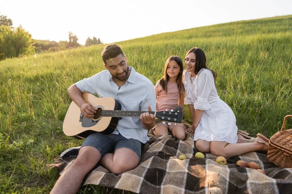 Homme jouant de la guitare à la famille heureuse pendant le pique-nique dans un champ herbeux — Photo de stock