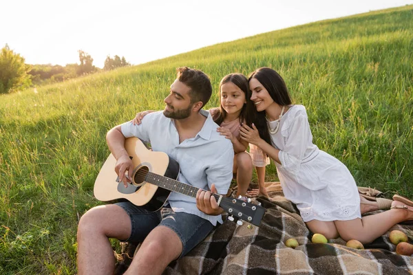 Весёлая женщина и ребёнок рядом с мужчиной играют на гитаре на пикнике в сельской местности — стоковое фото