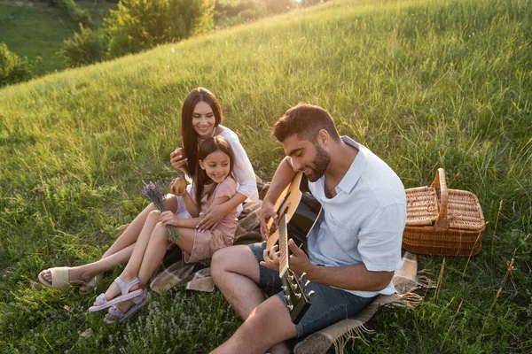 Homem tocando guitarra acústica para esposa feliz e filha em colina verde no dia de verão — Fotografia de Stock