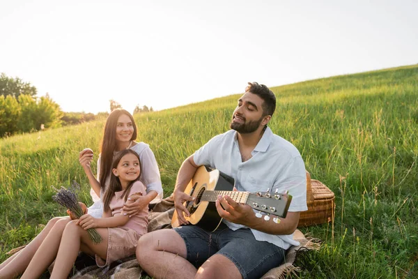 Pleased woman smiling near daughter and husband playing guitar outdoors - foto de stock