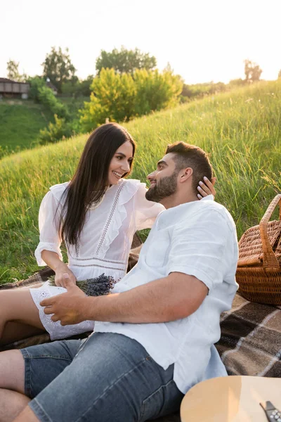 Brunette woman in white dress smiling at bearded man on picnic in countryside - foto de stock