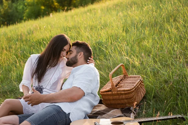 Glückliches Paar mit geschlossenen Augen, das sich auf dem Rasen neben Akustikgitarre und Weidenkorb umarmt — Stockfoto