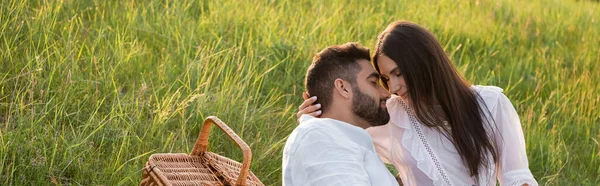 Brünette Frau mit geschlossenen Augen umarmt bärtigen Mann auf Rasen in der Nähe Weidenkorb, Banner — Stockfoto