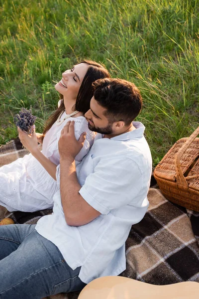 Smiling woman with closed eyes holding lavender bouquet near man hugging her during picnic — Foto stock