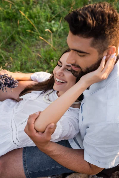 Brunette woman with lavender flowers touching face of bearded boyfriend while resting on lawn - foto de stock