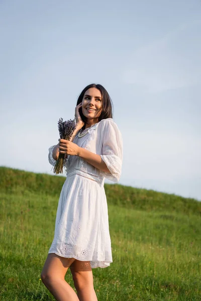Happy woman in white dress touching face and looking away in meadow — Fotografia de Stock