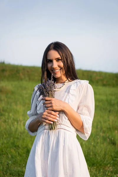 Morena mujer en vestido blanco sosteniendo ramo de lavanda y sonriendo a la cámara al aire libre - foto de stock