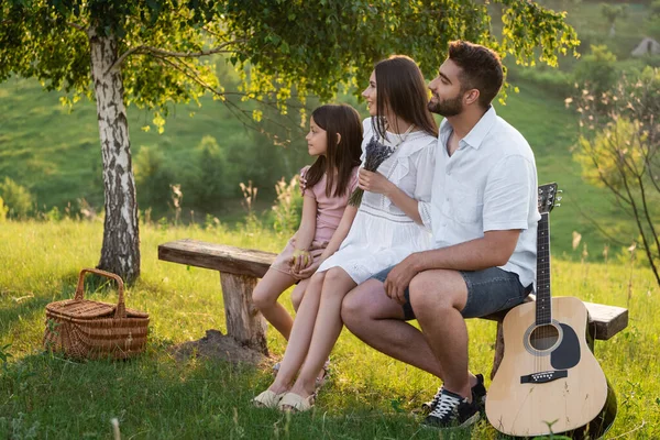 Улыбающаяся семья смотрит в сторону, сидя на скамейке под березой рядом с гитарой и плетеной корзиной — стоковое фото