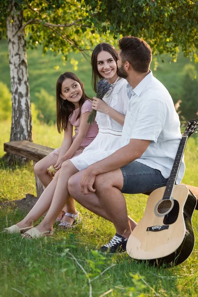 Glückliche Familie plaudert an einem Sommertag auf Bank neben Akustikgitarre — Stockfoto