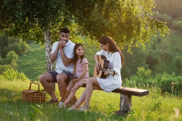 Couple playing harmonica and guitar to daughter on bench under birch — Fotografia de Stock