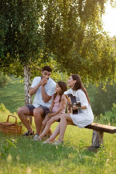Happy girl sitting on bench under birch near parents playing harmonica and guitar — Stock Photo