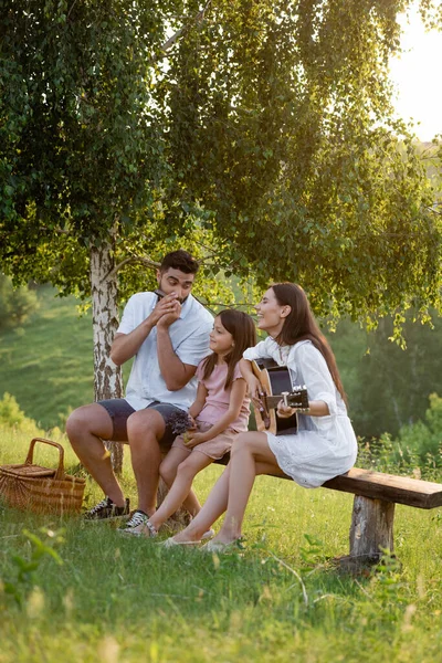 Ragazza felice seduta sulla panchina sotto betulla vicino ai genitori che suonano armonica e chitarra — Foto stock