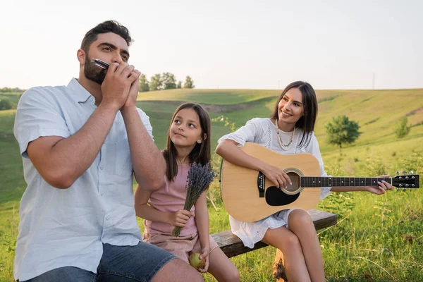 Hombre jugando armónica cerca de la esposa con la guitarra y el niño feliz en el campo - foto de stock