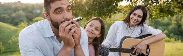 Man playing harmonica near cheerful daughter and wife with acoustic guitar, banner — Photo de stock