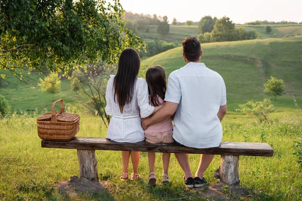 Задний вид семьи, сидящей на скамейке возле плетеной корзины спереди на живописном пейзаже — стоковое фото