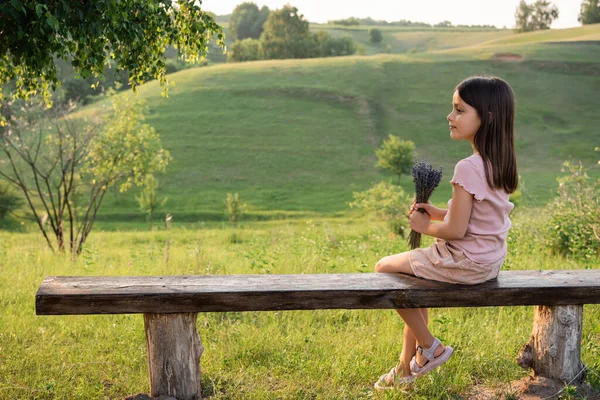 Vue latérale de la fille avec des fleurs de lavande assis sur le banc près des collines pittoresques — Photo de stock