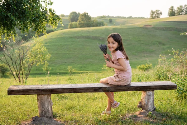 Cheerful girl with lavender flowers sitting on bench in meadow and looking at camera - foto de stock