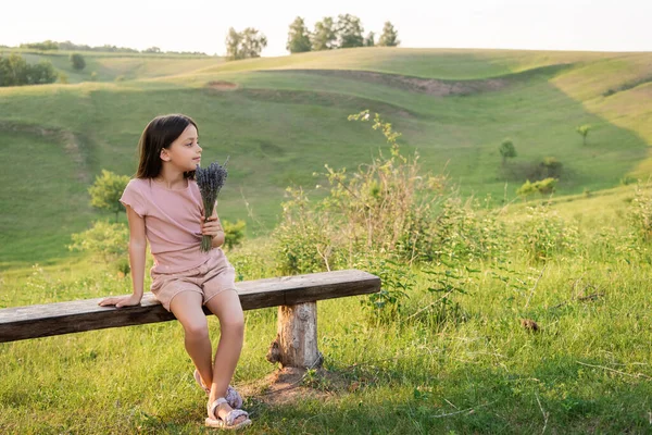 Full length of girl with lavender flowers sitting on bench in countryside and looking away — Fotografia de Stock