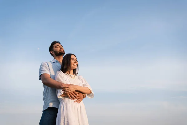 Bearded man embracing happy woman in white dress while looking away under blue sky - foto de stock