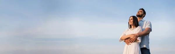 Joyful man hugging brunette woman in white dress and looking away under clear sky, banner — Stockfoto