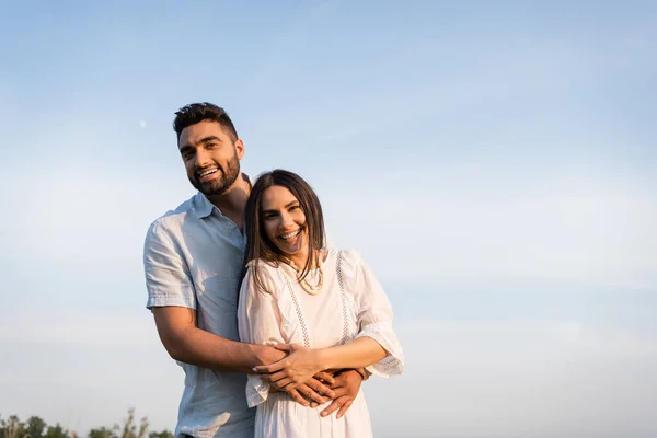 Homme joyeux étreignant femme brune en robe blanche et regardant la caméra à l'extérieur — Photo de stock