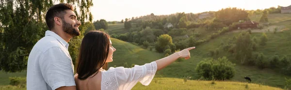 Femme souriante pointant vers les collines pittoresques près de l'homme heureux à la campagne, bannière — Photo de stock